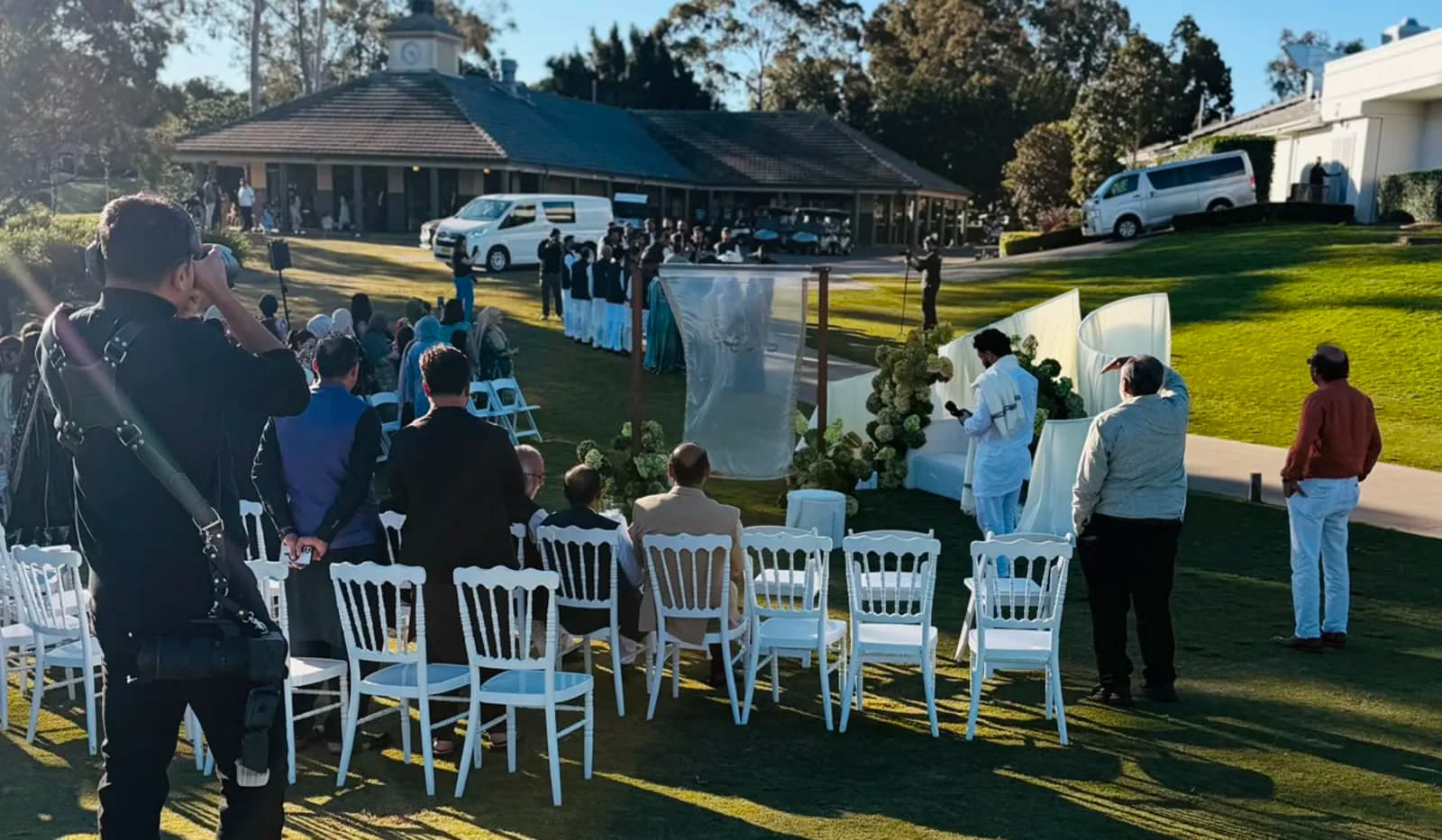 An outdoor ceremony under way on the lawn, with guests seated in white chairs